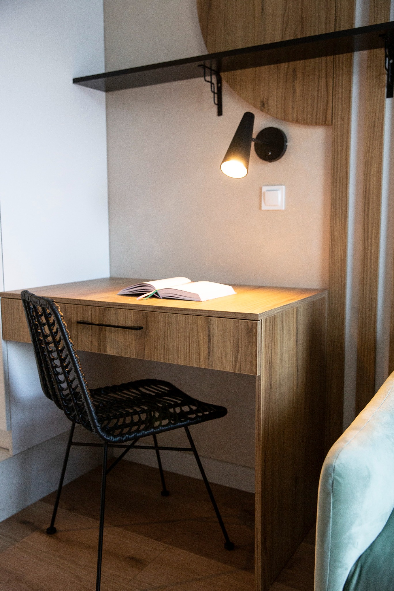 Drewniane biurko z czarnym krzesłem i otwartą książką, oświetlone lampką ścienną. Minimalistyczny design, ciepłe oświetlenie, fragment sofy w tle.