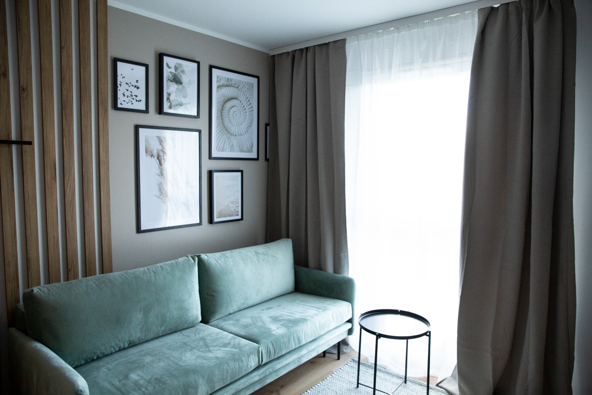 Pastelowa sofa w pokoju z galerią obrazów i jasnymi zasłonami. Minimalistyczny stolik kawowy i drewniane lamele dodają wnętrzu nowoczesnego charakteru.