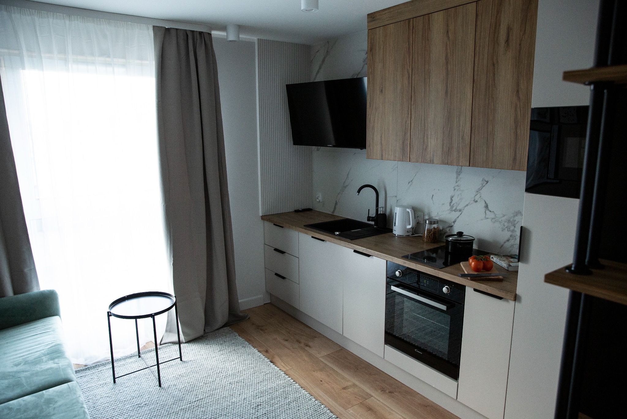 Jasna kuchnia z białymi szafkami, drewnianym blatem i marmurową ścianą. Czarny zlewozmywak i piekarnik. W tle okno z zasłonami i fragment salonu.