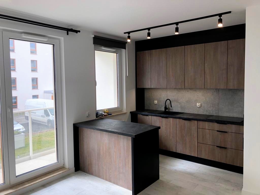 Nowoczesna kuchnia z drewnianymi frontami, czarnym blatem i oświetleniem punktowym. Widok na balkon i budynek z okna. Minimalistyczny design.