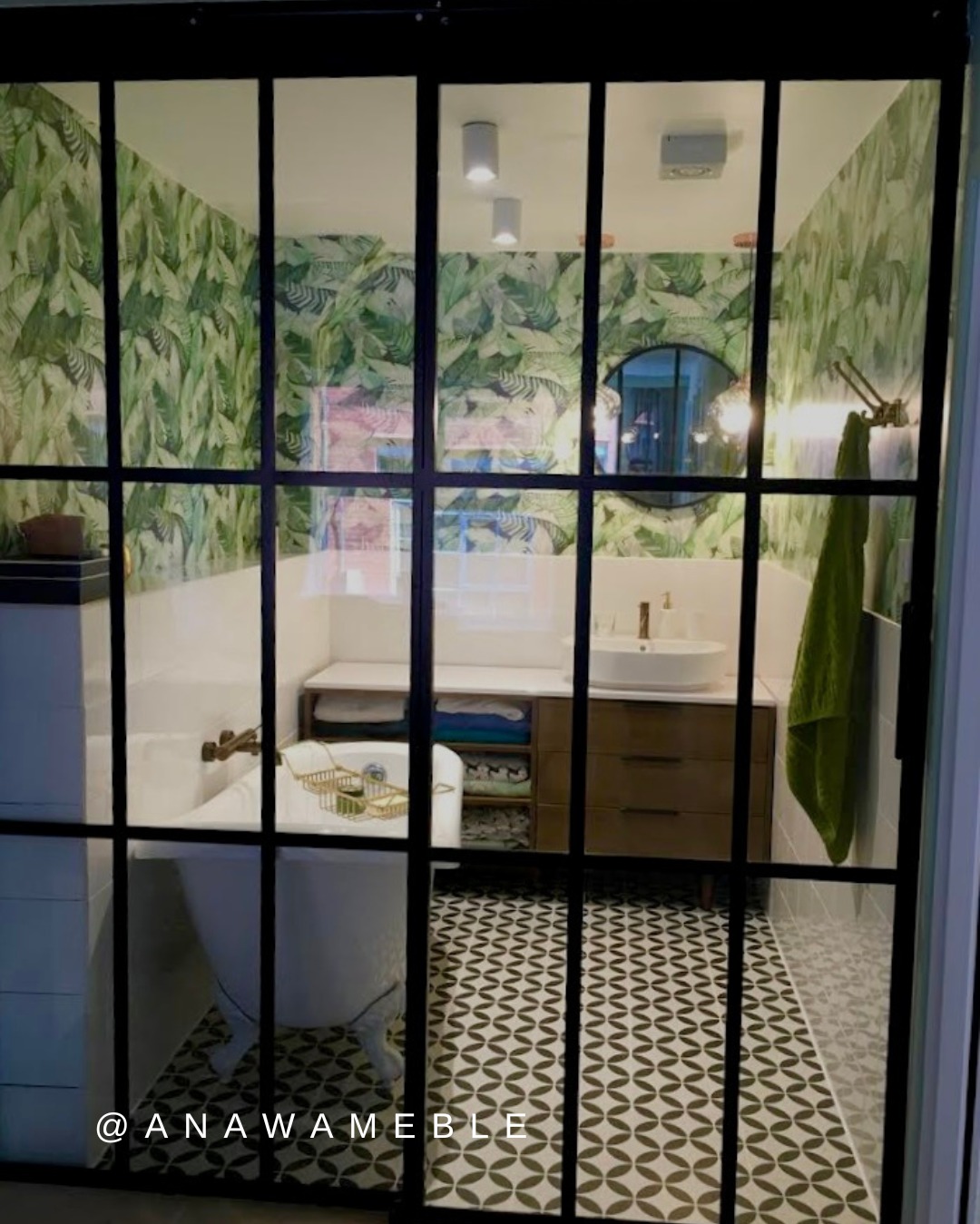 Stylowa łazienka za czarną, szklaną ścianą z podziałami. Wanna na nóżkach, wzorzysta podłoga, ściany w liście. Nowoczesne oświetlenie i okrągłe lustro dopełniają aranżację.