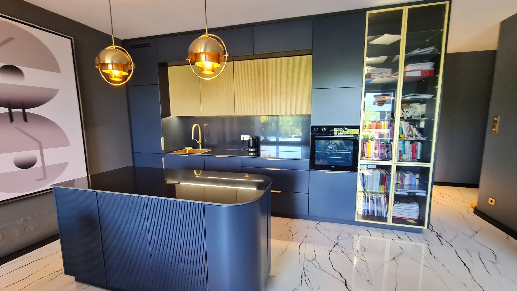Nowoczesna kuchnia w odcieniach granatu i złota, z wyspą i marmurową podłogą. Widoczne lampy wiszące i przeszklona witryna.