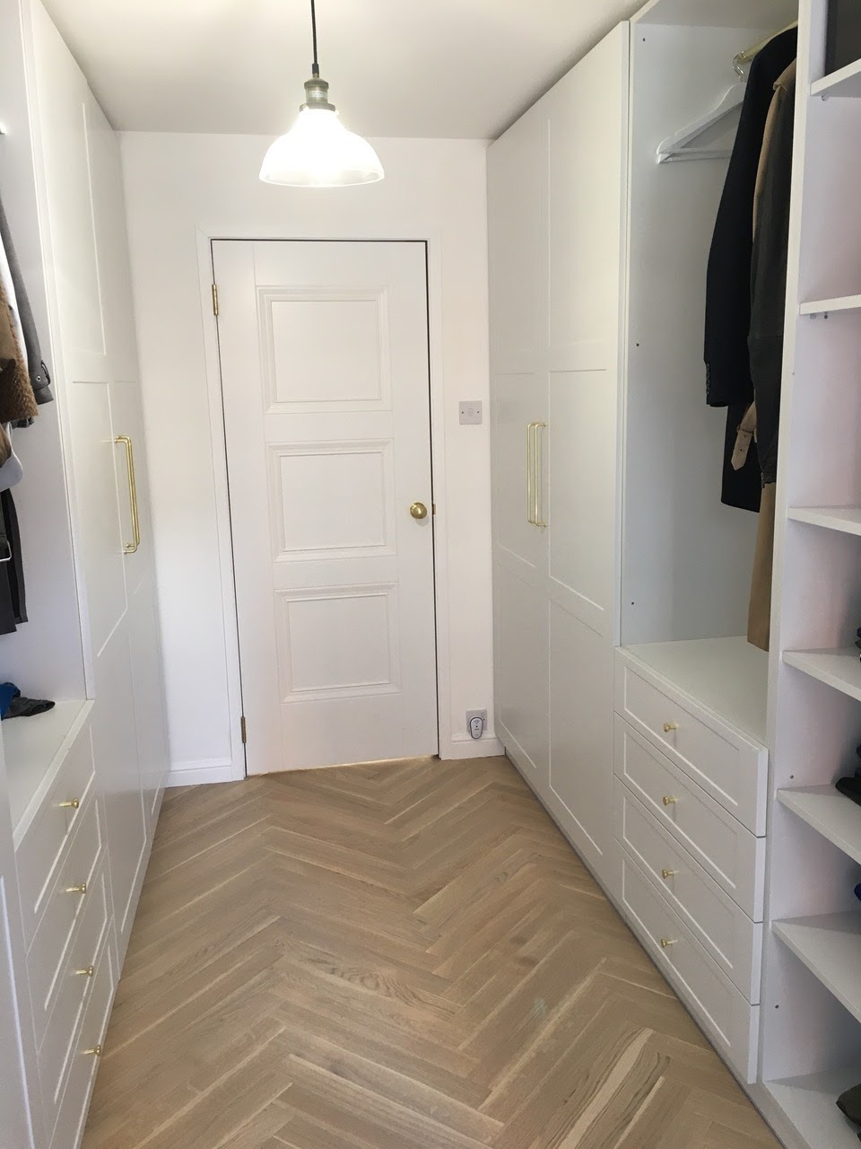 Biała garderoba z parkietem ułożonym w jodełkę, oświetlona lampą wiszącą. Minimalistyczny design, złote uchwyty, drzwi w tle. Widok na wnętrze szafy z ubraniami.