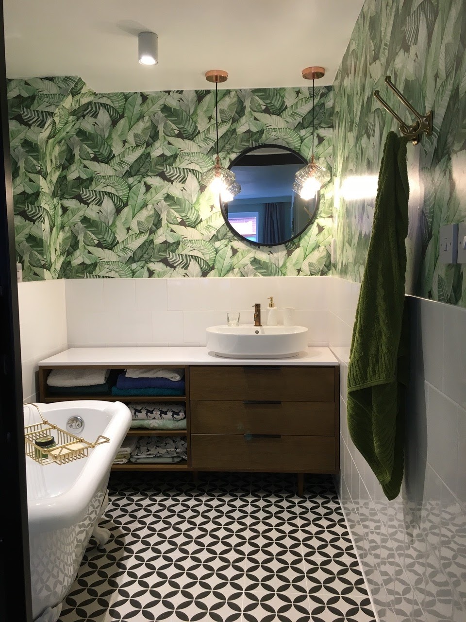 Stylowa łazienka z wanną na nóżkach, umywalką nablatową i wzorzystą tapetą w liście. Uwagę przykuwają lampy wiszące z miedzianymi elementami i geometryczna podłoga.