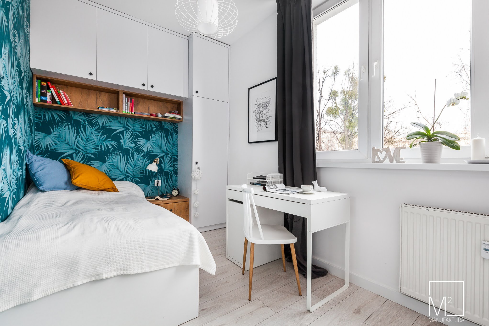 Jasna sypialnia z turkusową tapetą w liście, białymi meblami i drewnianymi akcentami. Widok na łóżko, biurko z krzesłem i okno z dekoracjami.