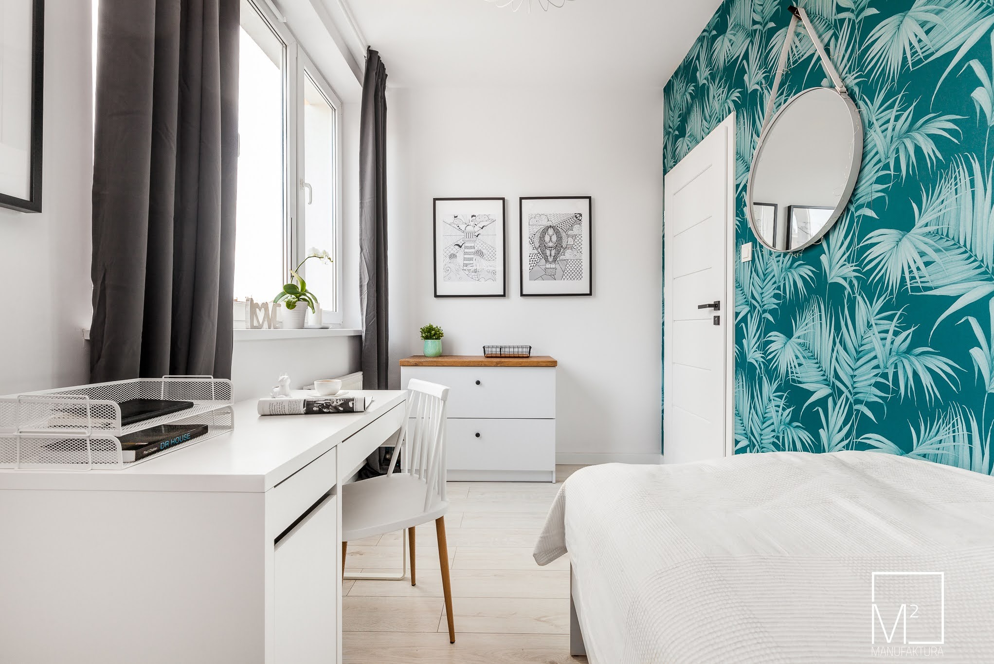 Jasne wnętrze sypialni z biurkiem, białymi meblami i turkusową tapetą w liście. Nad komodą wiszą czarno-białe grafiki w ramkach, a nad łóżkiem okrągłe lustro.
