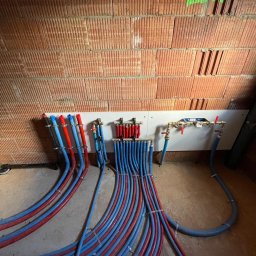 Przemysław Grzegorczyk - Rozdzielacz ogrzewania podłogowego z rurami PEX w kolorach niebieskim i czerwonym, zamocowany na ścianie z cegły dziurawki. Instalacja w surowym stanie.