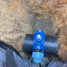 Przemysław Grzegorczyk - Niebieski zawór na czarnej rurze w wykopie z piaskiem, widok z góry,  element instalacji wodociągowej w ziemi, robocze spodnie i buty w kadrze.