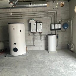 Przemysław Grzegorczyk - Biały bojler Vaillant i dwa piece gazowe na tle ściany z szarych płytek, z widoczną instalacją rur. Całość w surowym wnętrzu z betonową podłogą i sufitem.