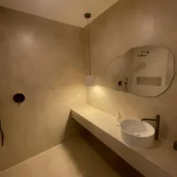 Nowoczesna łazienka w beżowych tonacjach: umywalka nablatowa, minimalistyczna armatura, asymetryczne lustro i oświetlenie punktowe. Widoczny fragment kabiny prysznicowej.