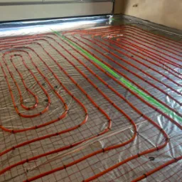 Pomarańczowe rury ogrzewania podłogowego ułożone na srebrnej folii z siatką, zabezpieczone czarnymi klipsami. Widoczny zielony laser wyznaczający poziom.