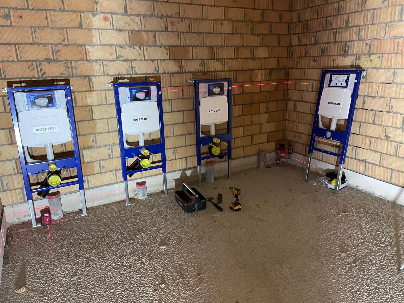 Montaż stelaży podtynkowych Geberit w surowym wnętrzu z ceglanymi ścianami. Laserowy niwelator i narzędzia hydrauliczne widoczne na podłodze.
