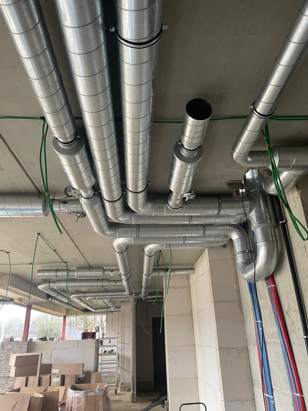 Widok na sufit z zamontowanymi, srebrnymi rurami wentylacyjnymi, przymocowanymi do betonowego stropu. Widoczne kable i fragment ściany z instalacjami.