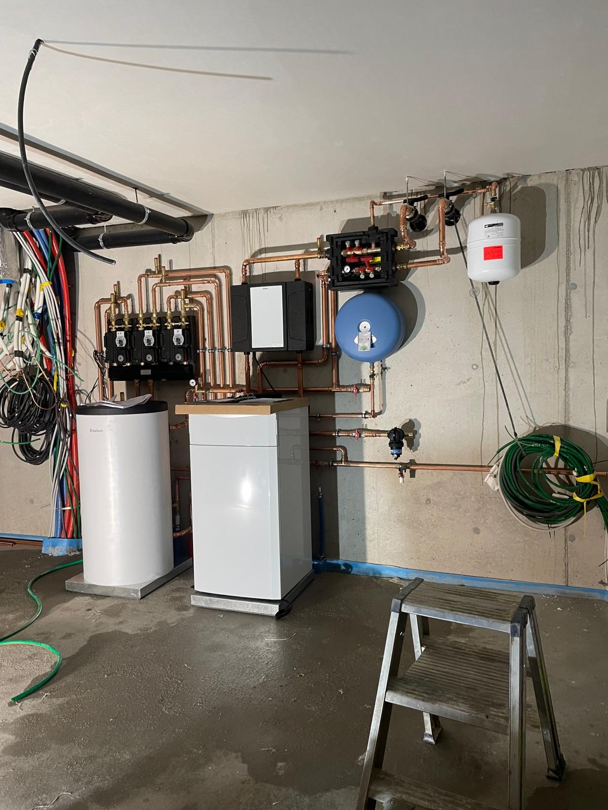 Kompleksowa instalacja grzewcza z dwoma białymi piecami Vaillant, miedzianymi rurami i czarną skrzynką rozdzielczą na szarej betonowej ścianie. Na pierwszym planie drabina.