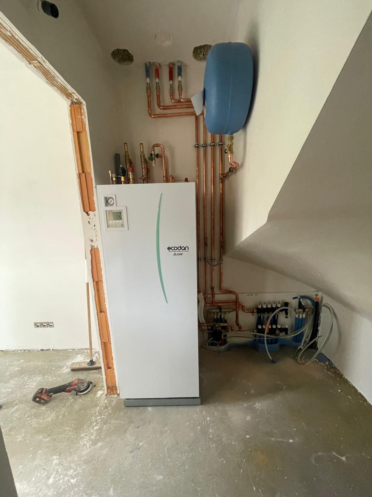 Biały piec gazowy Ecodan Mitsubishi z miedzianą instalacją, zbiornikiem wyrównawczym i rozdzielaczem w surowym pomieszczeniu, widoczne narzędzie szlifierskie.