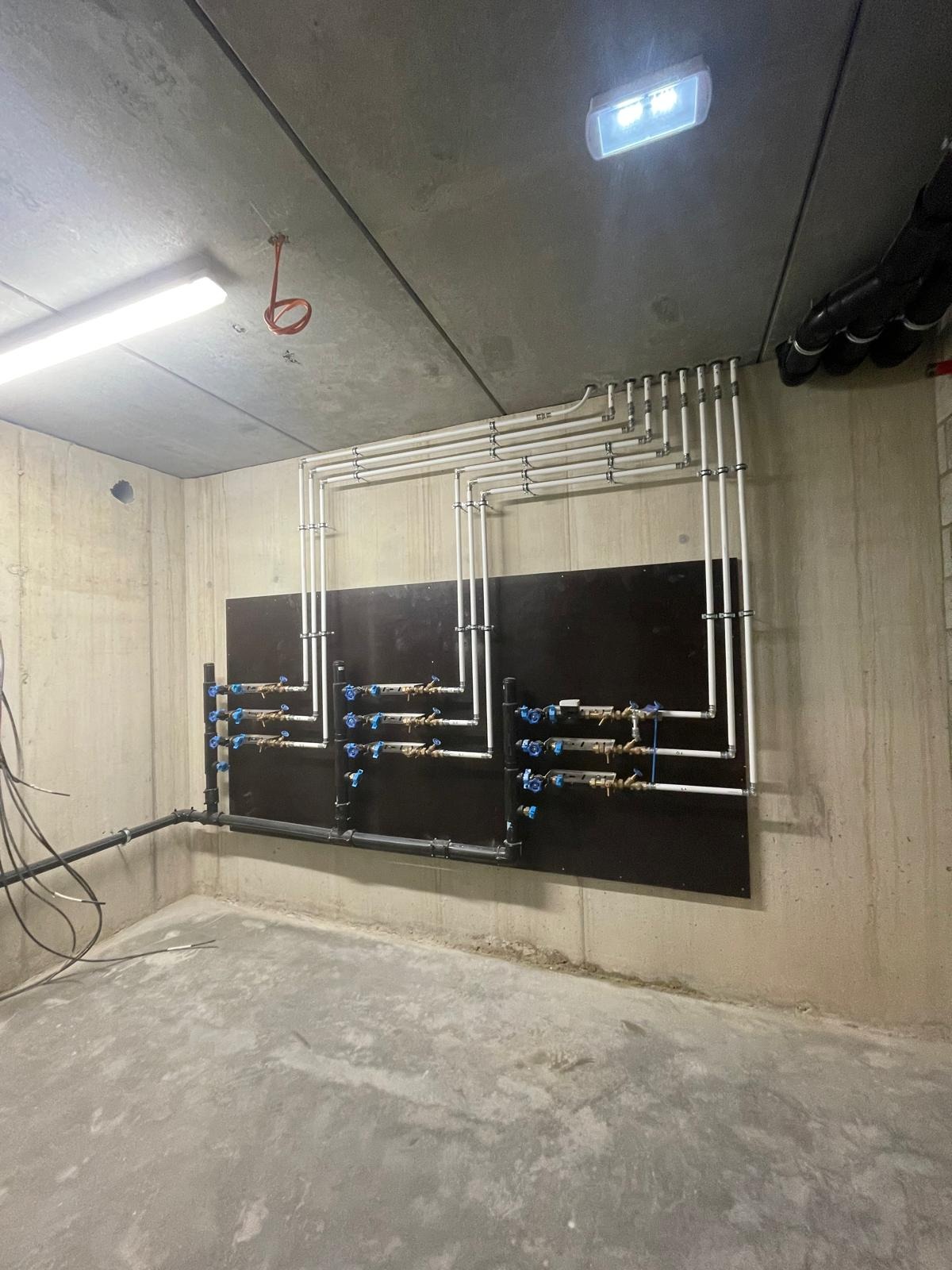 Instalacja wodna z licznymi zaworami i rurami w surowym pomieszczeniu z betonowymi ścianami i podłogą. Rury przymocowane do czarnej płyty na ścianie.
