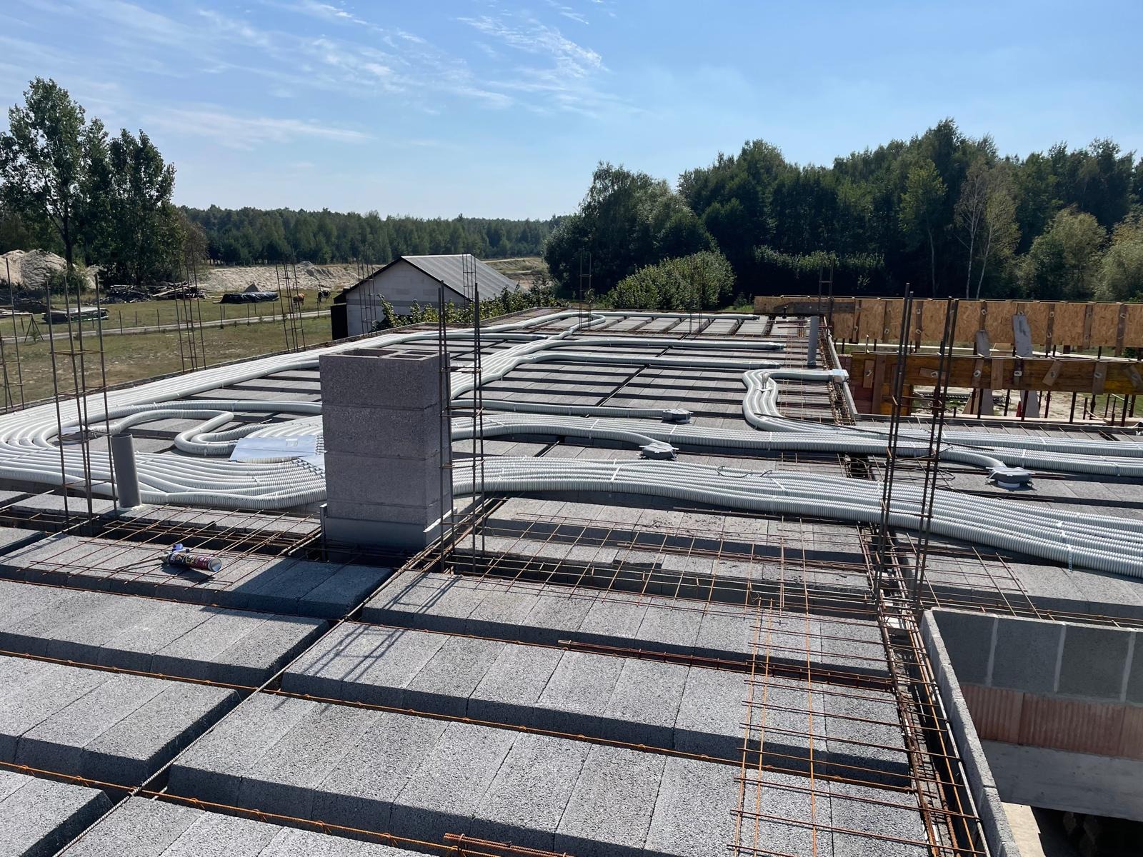 Szare bloczki stropowe z rozłożoną instalacją rekuperacji w białych rurach karbowanych. Widoczne zbrojenie, komin z bloczków i fragment lasu w tle.