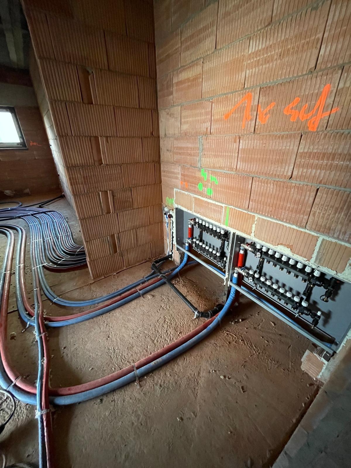 Rozdzielacze ogrzewania podłogowego zamontowane w ścianie z cegły, z widocznymi rurami doprowadzającymi. Surowy stan budynku.