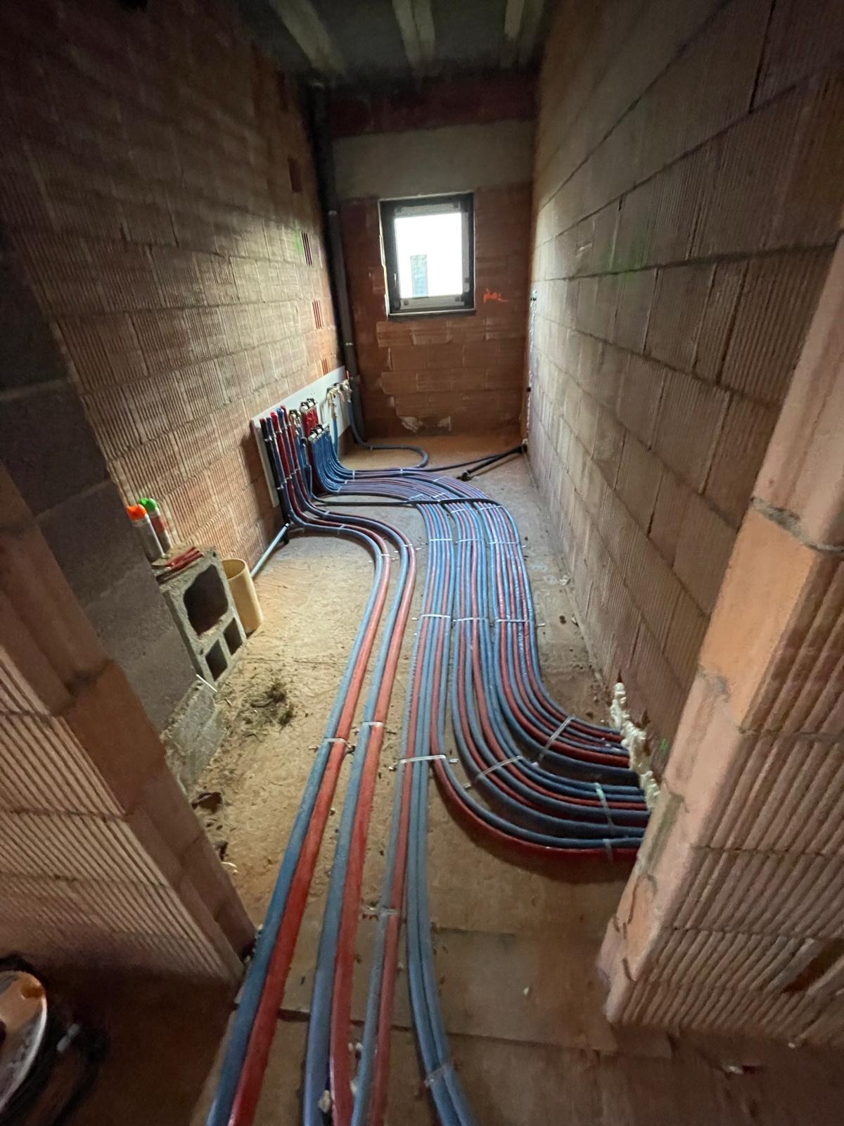 Instalacja rur grzewczych (czerwone i niebieskie) ułożonych wzdłuż ściany z cegły w surowym wnętrzu budynku, widok z góry, etap prac instalacyjnych.