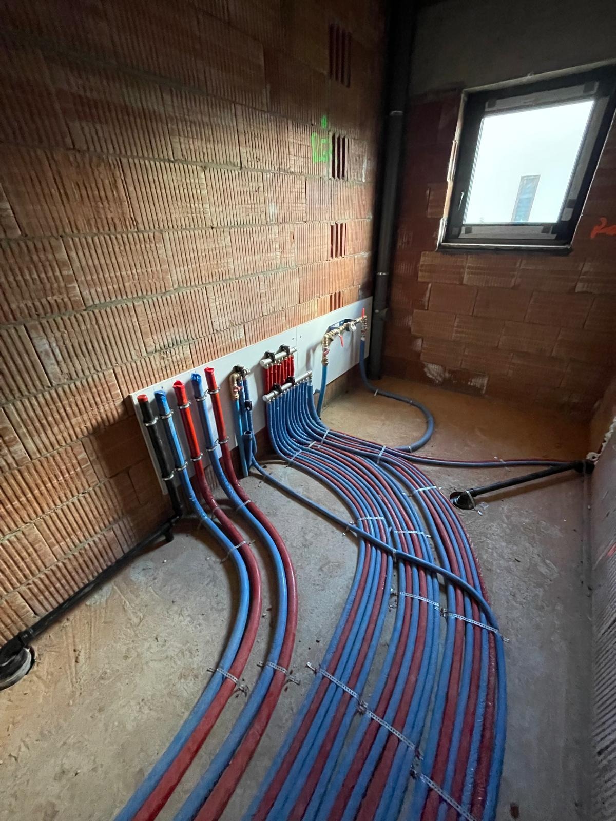 Rozprowadzenie instalacji wodnej z rur PEX w kolorach niebieskim i czerwonym, zamocowanych do ściany z cegły, w surowym wnętrzu z oknem.