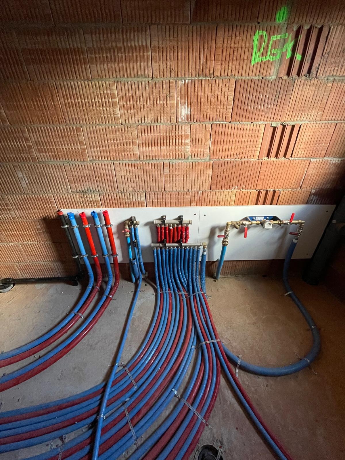 Rozdzielacz ogrzewania podłogowego z rurami PEX w kolorach niebieskim i czerwonym, zamocowany na ścianie z cegły dziurawki. Instalacja w surowym stanie.