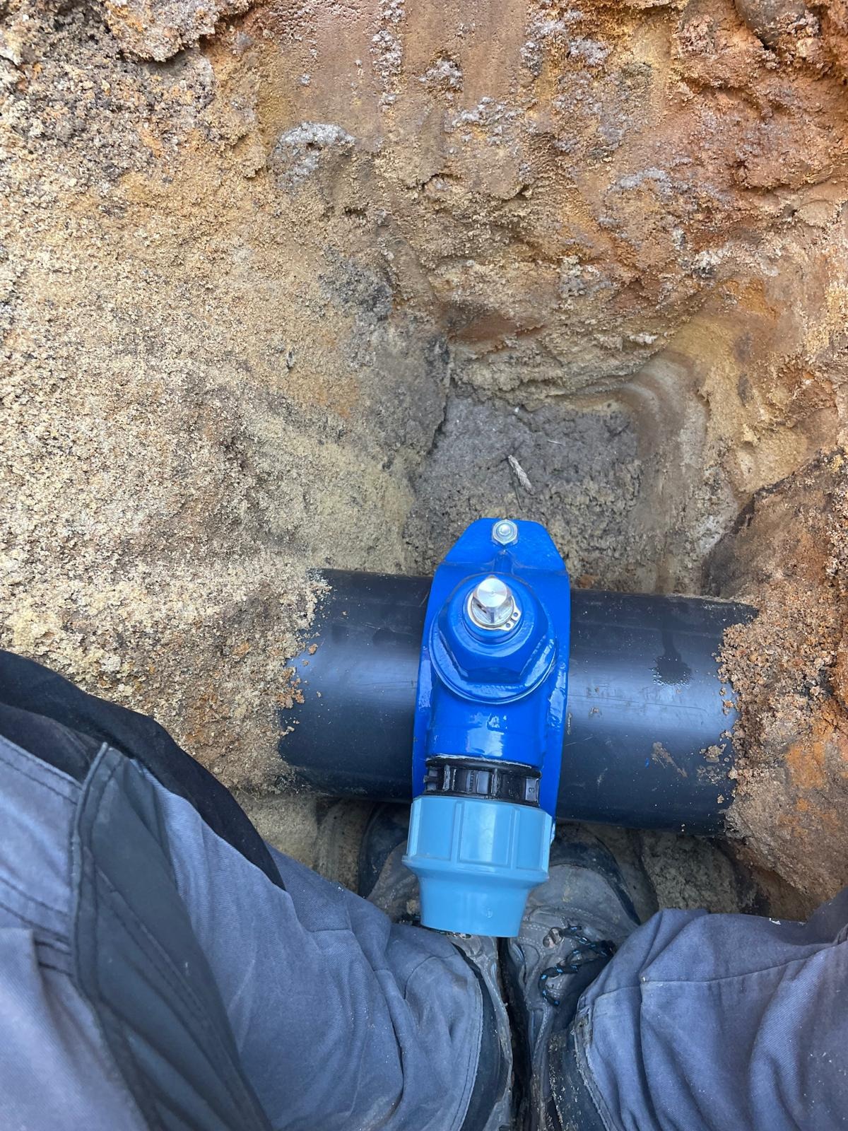 Niebieski zawór na czarnej rurze w wykopie z piaskiem, widok z góry,  element instalacji wodociągowej w ziemi, robocze spodnie i buty w kadrze.