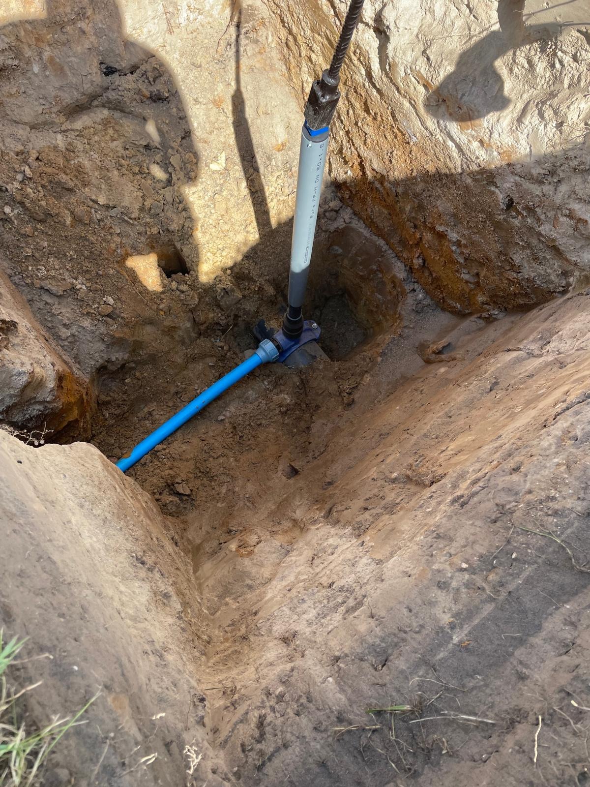 Nowe przyłącze wodociągowe w wykopie ziemnym. Widoczne rury z tworzywa sztucznego i elementy złączek. Świeże wykopy i cień osoby wykonującej pracę.