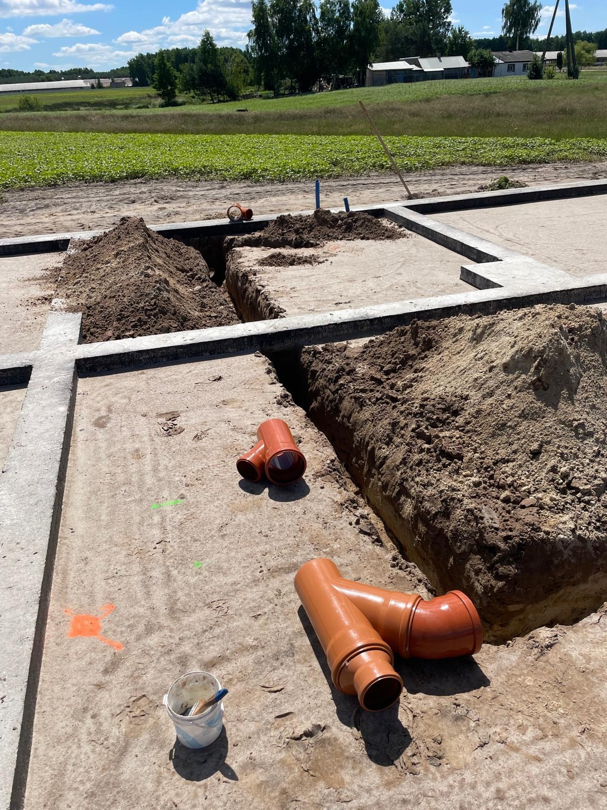 Wykonane wykopy pod fundamenty z widocznymi pomarańczowymi rurami kanalizacyjnymi, przygotowane do instalacji. Widok na pole i zabudowania w tle.