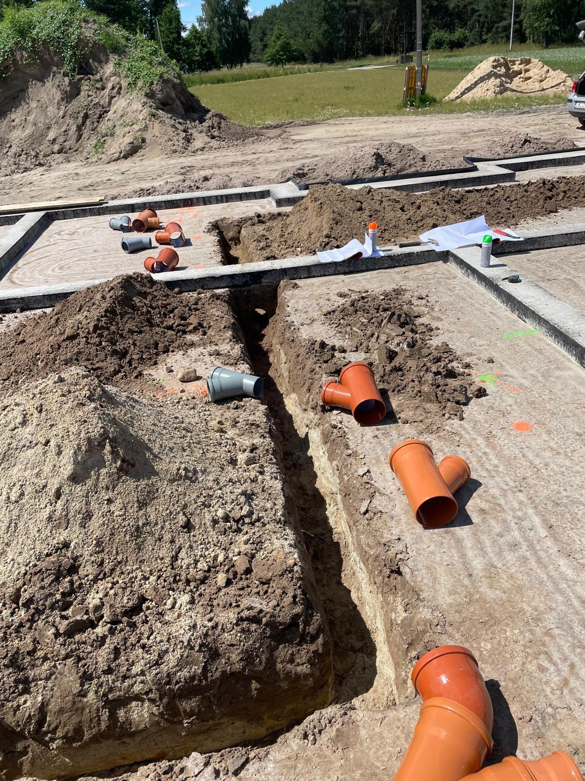 Wykop pod fundamenty z ułożonymi rurami kanalizacyjnymi i drenażowymi, widoczne złącza Y. Na ziemi leżą plany, a w tle widać drzewa i niebo.