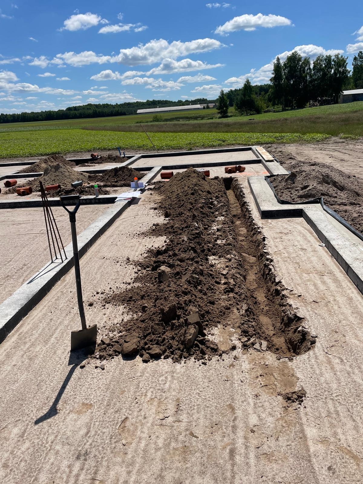 Wykop pod fundamenty z ułożonymi rurami kanalizacyjnymi na tle zielonego pola i błękitnego nieba. Widoczna łopata i inne narzędzia.