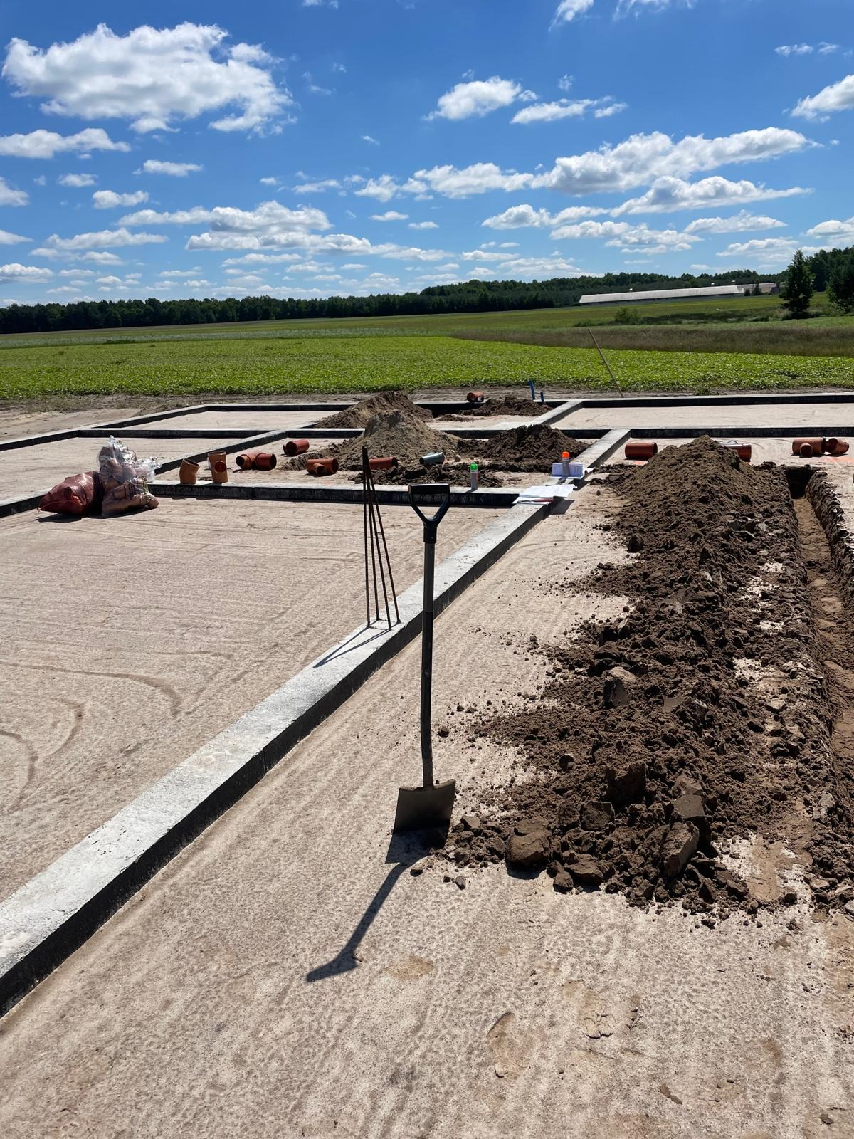 Fundamenty budynku z przygotowanymi wykopami pod instalacje, widoczne rury i narzędzia. Prace ziemne w toku, słoneczny dzień, pola w tle.
