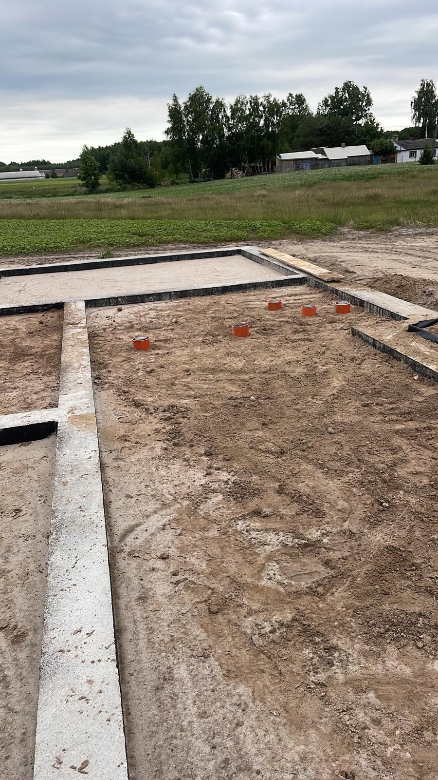 Fundamenty domu z wyprowadzonymi rurami kanalizacyjnymi, widok z góry na plac budowy w słoneczny dzień. Betonowe belki i pomarańczowe rury.