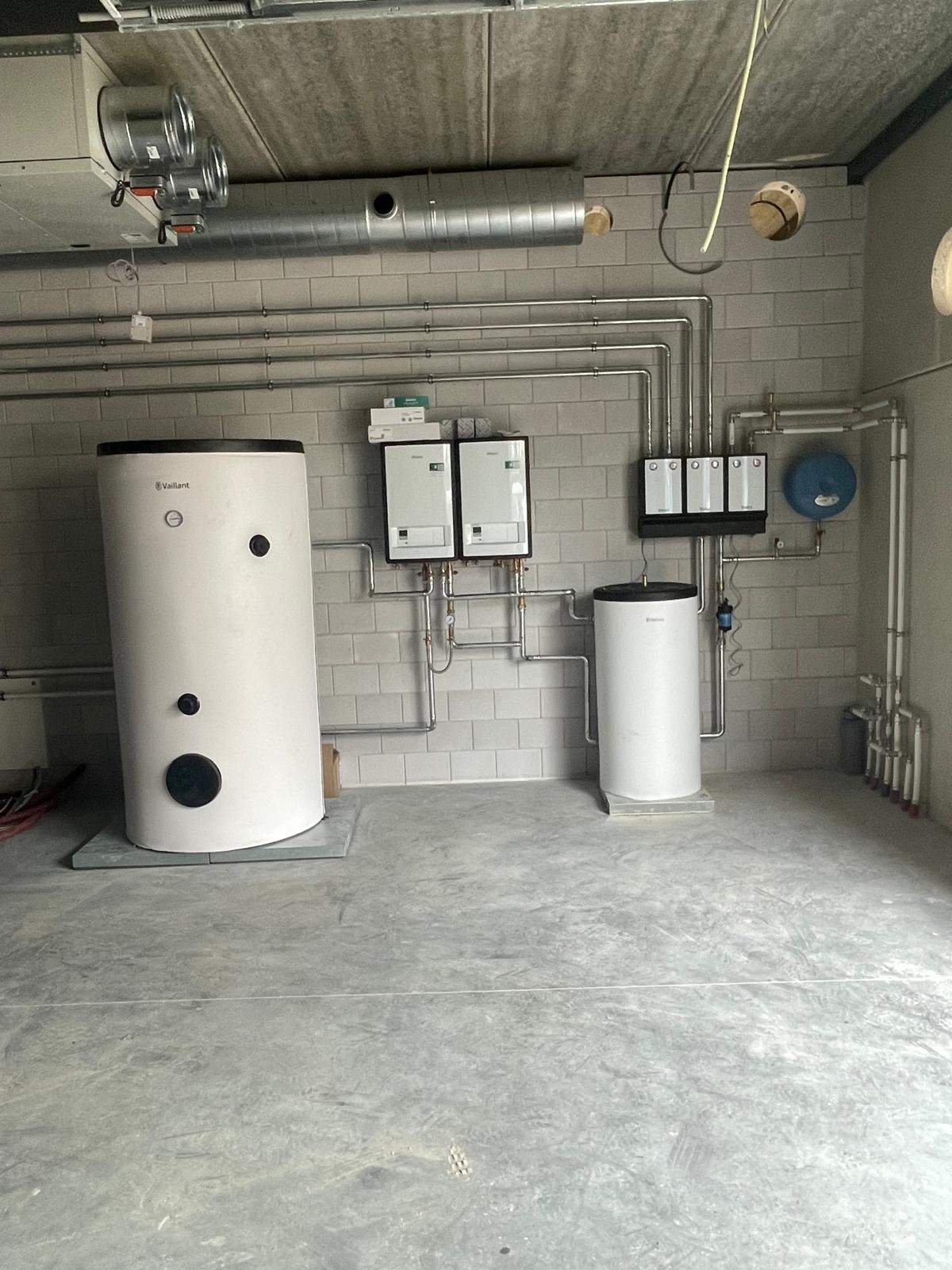 Biały bojler Vaillant i dwa piece gazowe na tle ściany z szarych płytek, z widoczną instalacją rur. Całość w surowym wnętrzu z betonową podłogą i sufitem.