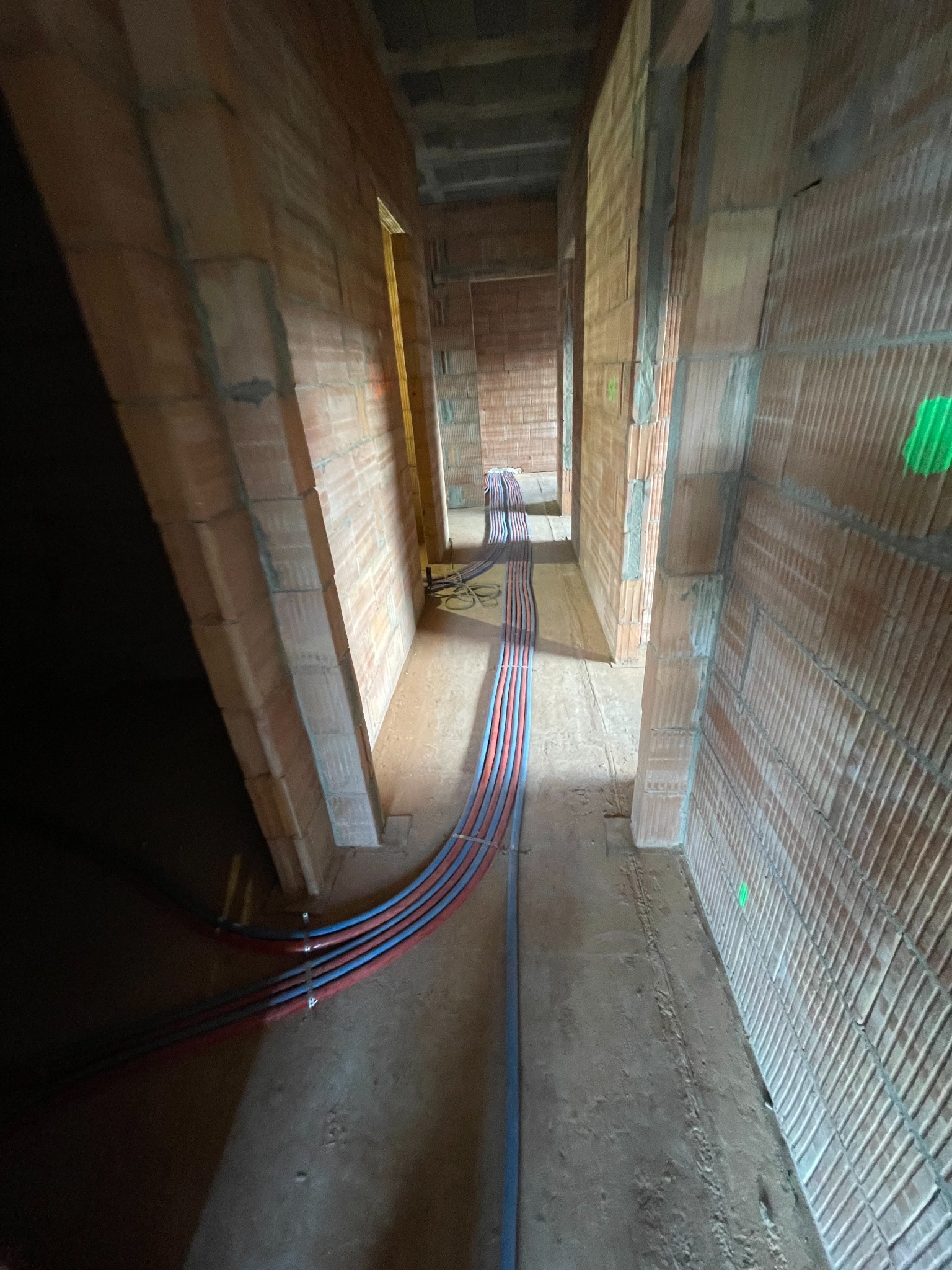Wnętrze w trakcie budowy z widocznymi przewodami instalacji grzewczej ułożonymi wzdłuż korytarza, ściany z cegły dziurawki, surowy stan.