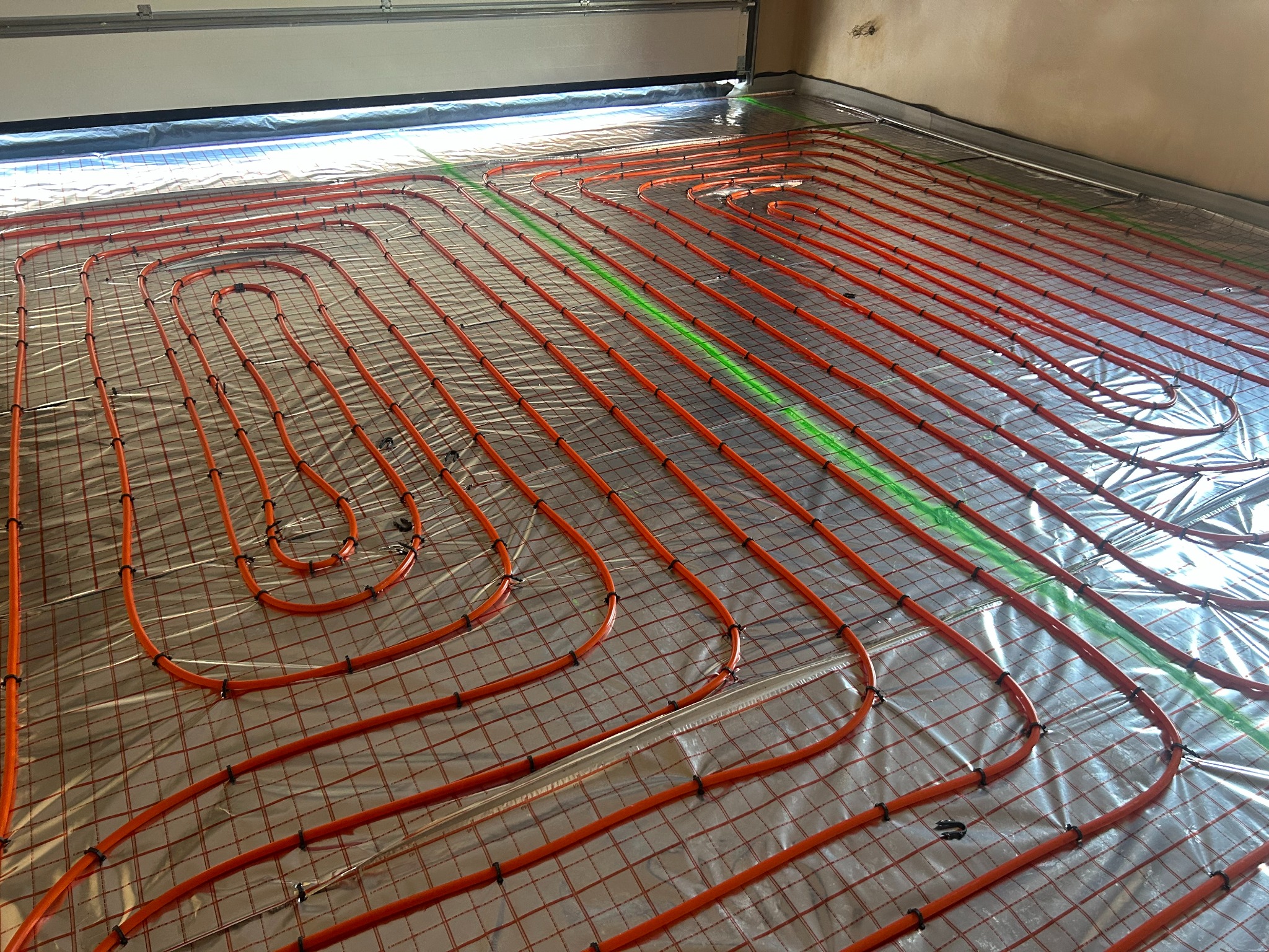 Pomarańczowe rury ogrzewania podłogowego ułożone na srebrnej folii z siatką, zabezpieczone czarnymi klipsami. Widoczny zielony laser wyznaczający poziom.