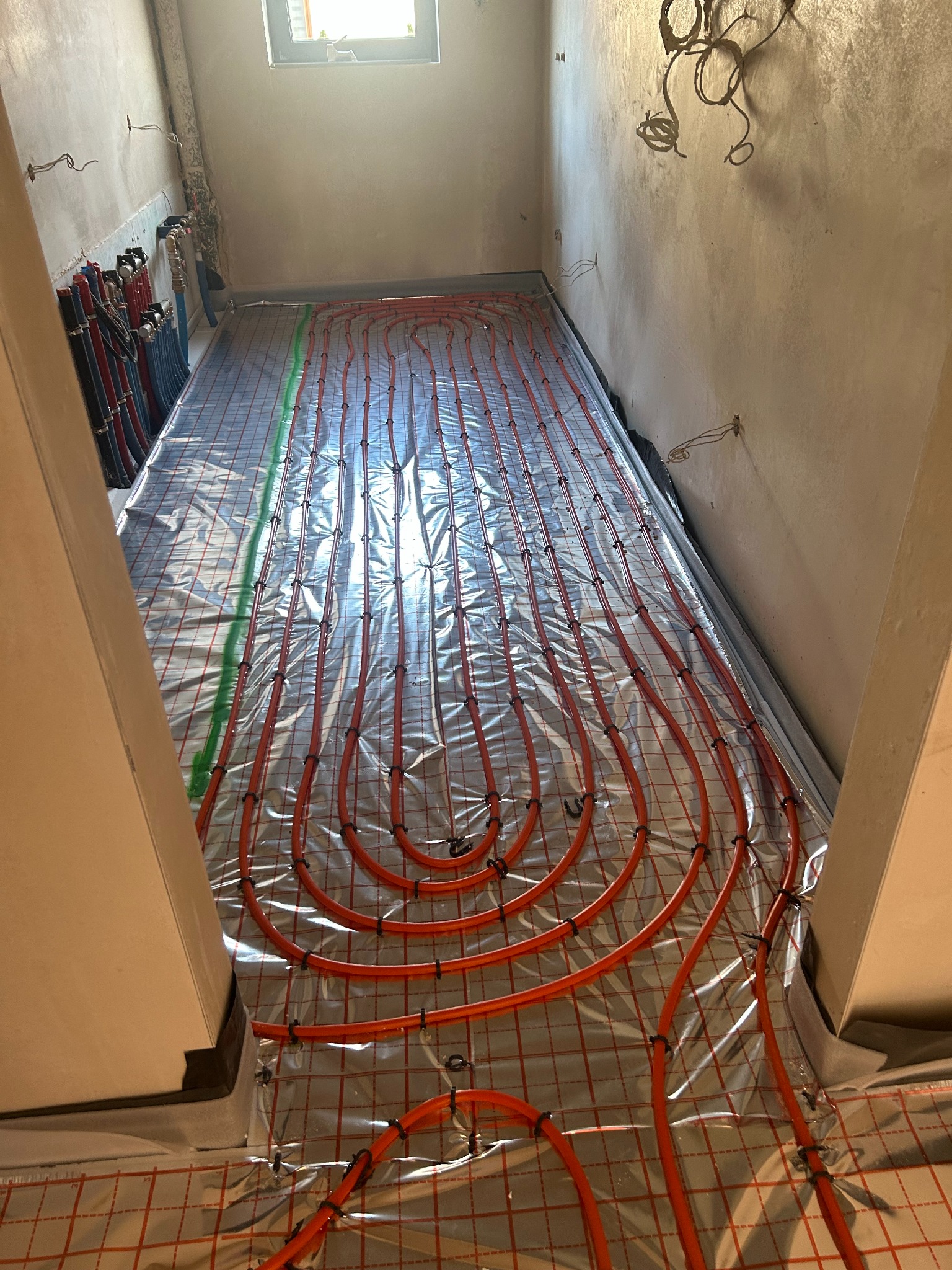 Układanie pomarańczowych rur ogrzewania podłogowego na folii z siatką w surowym wnętrzu. Widoczne pionowe rury przy ścianie i okno w górze.