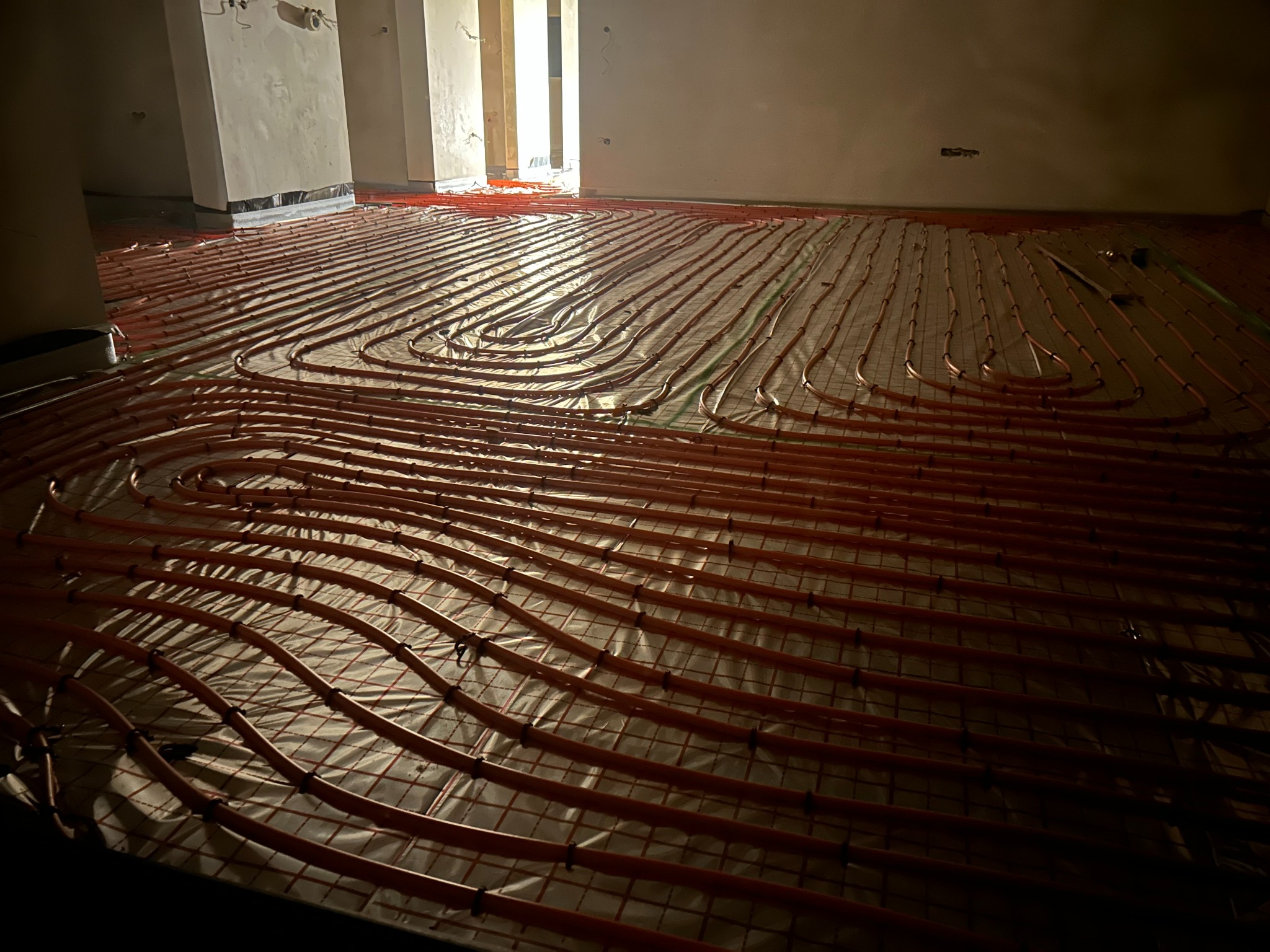 Rozłożona instalacja ogrzewania podłogowego z rurkami w odcieniach czerwieni na folii izolacyjnej w surowym wnętrzu. Widoczne fragmenty ścian i kolumn w tle.