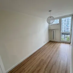 Jasne wnętrze pokoju po remoncie: drewnopodobna podłoga, kremowe ściany, białe listwy przypodłogowe i nowoczesna lampa wisząca.