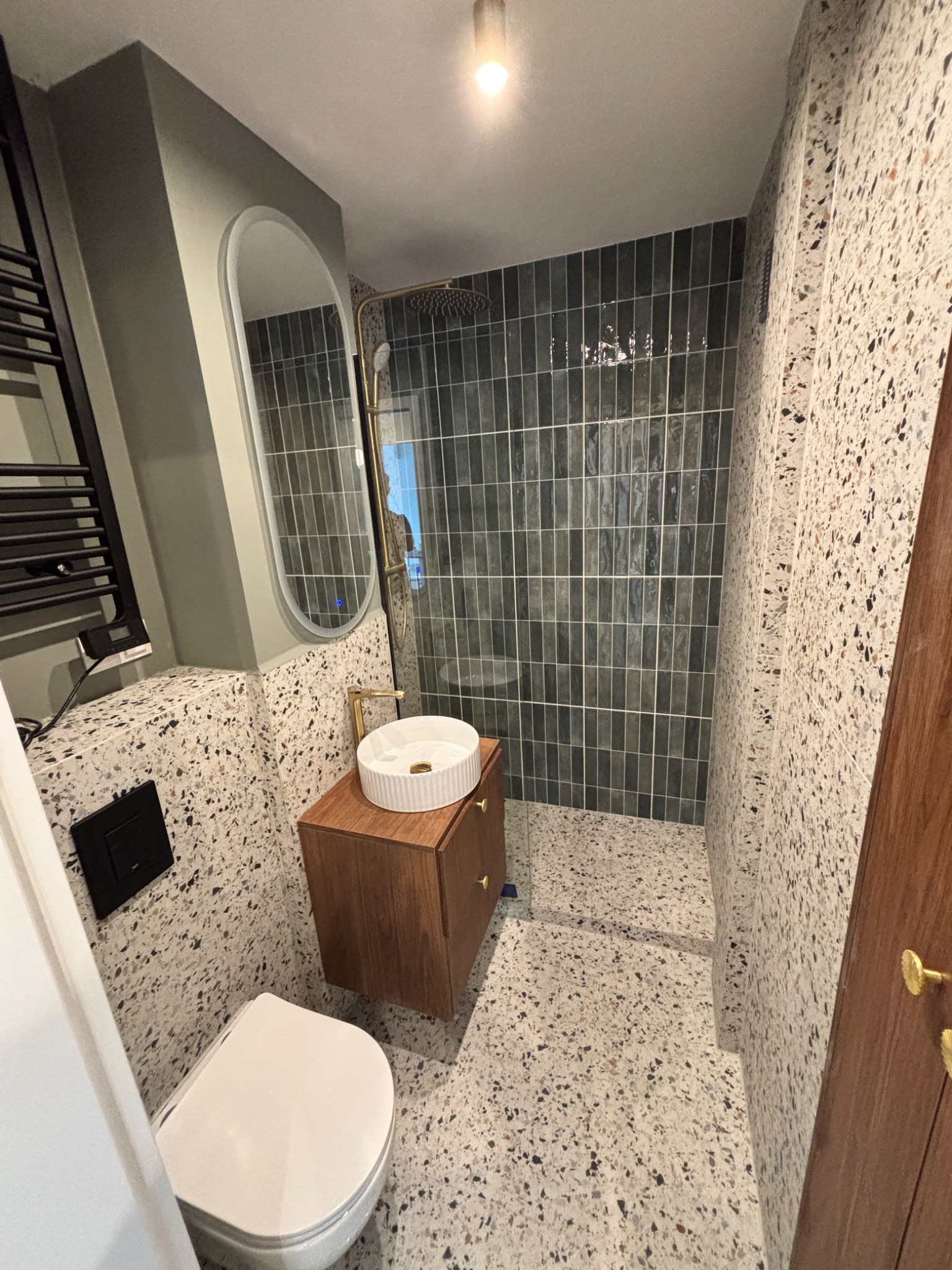 Nowoczesna łazienka z umywalką nablatową, kabiną prysznicową z ciemnymi płytkami i podłogą w stylu lastryko. Lustro w owalnej ramie i grzejnik drabinkowy.