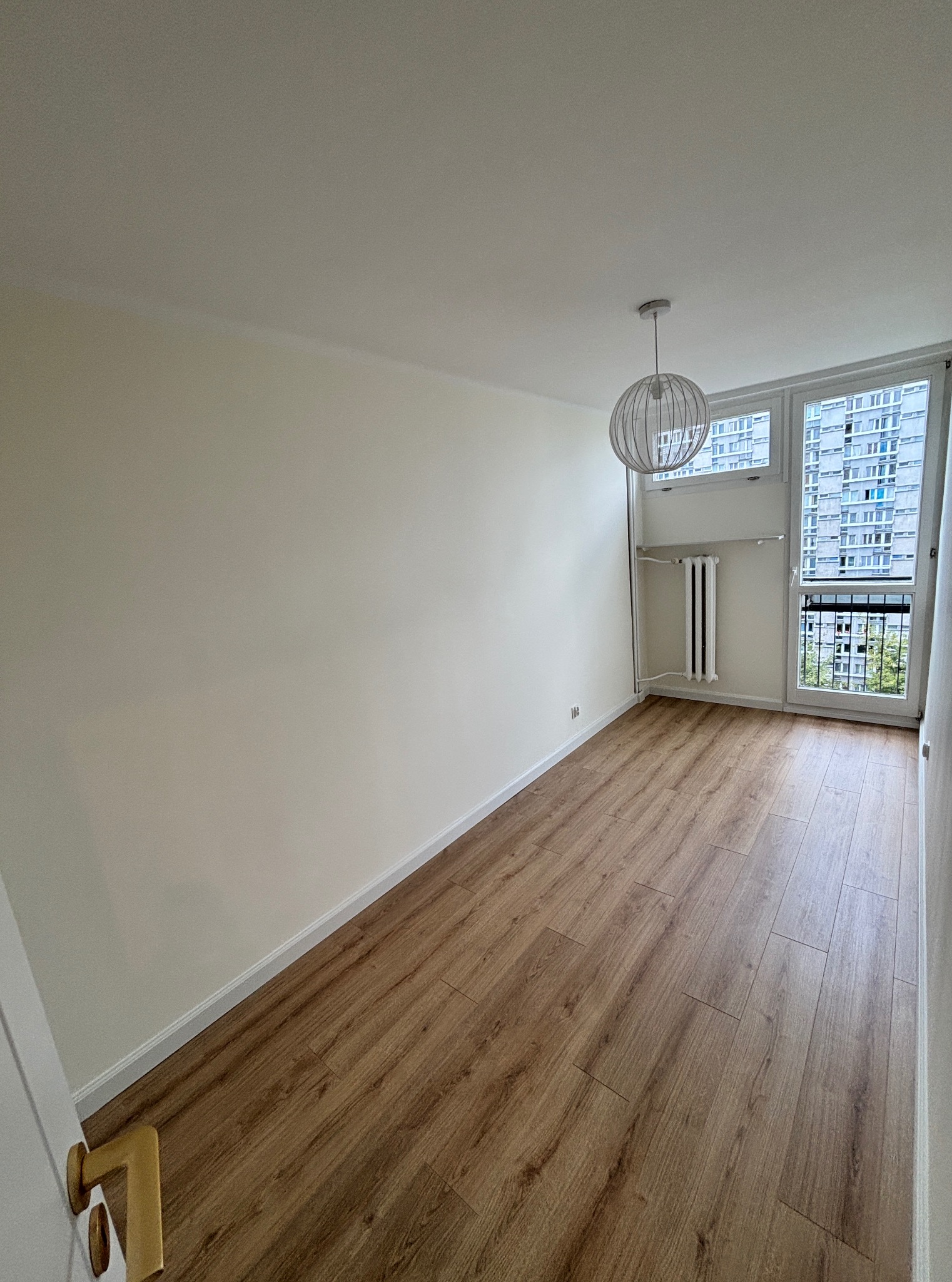 Jasne wnętrze pokoju po remoncie: drewnopodobna podłoga, kremowe ściany, białe listwy przypodłogowe i nowoczesna lampa wisząca.