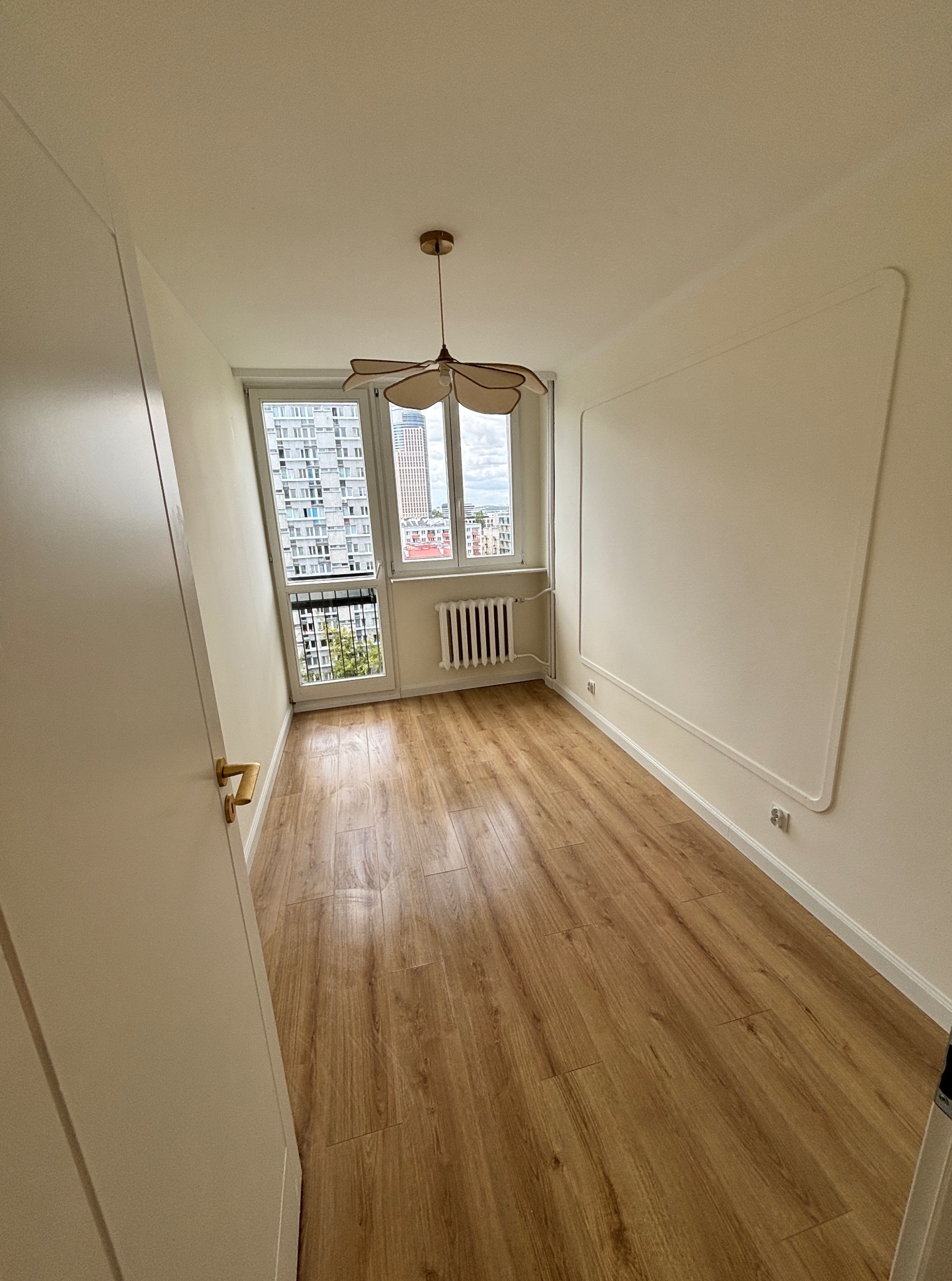 Jasne wnętrze pokoju po remoncie: drewniana podłoga, białe ściany z dekoracyjną ramą, okno z widokiem na miasto, designerska lampa sufitowa.