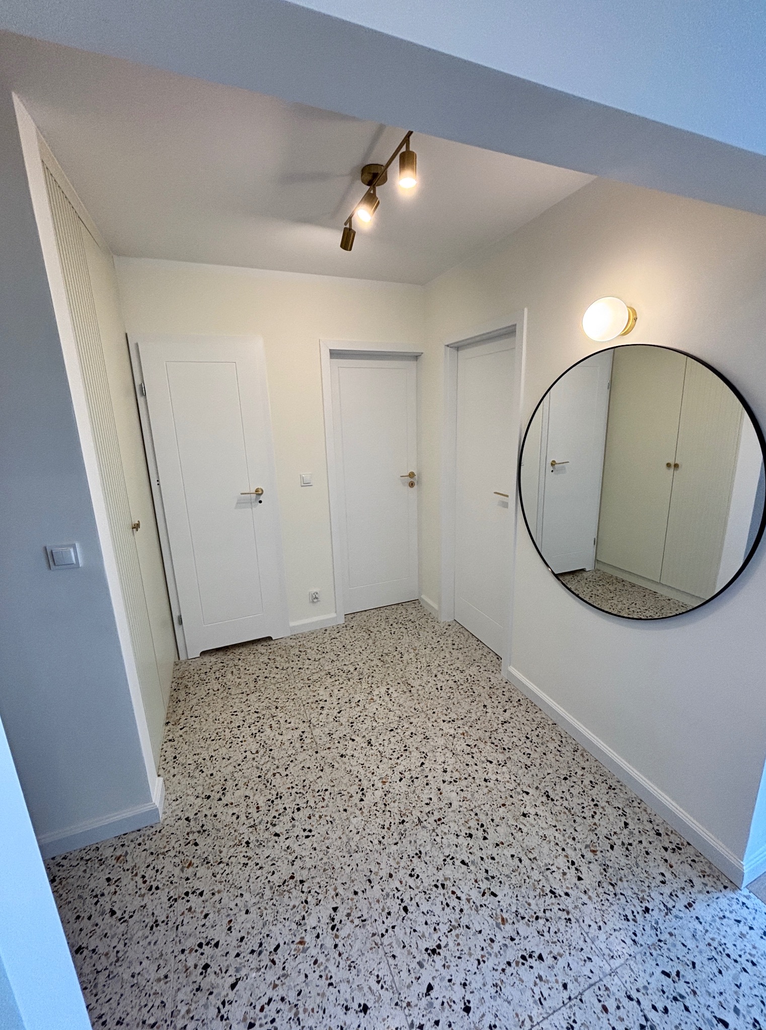 Jasne wnętrze przedpokoju z terazzo na podłodze, białymi drzwiami i okrągłym lustrem z czarną ramą. Oświetlenie sufitowe i ścienne w kolorze złota.