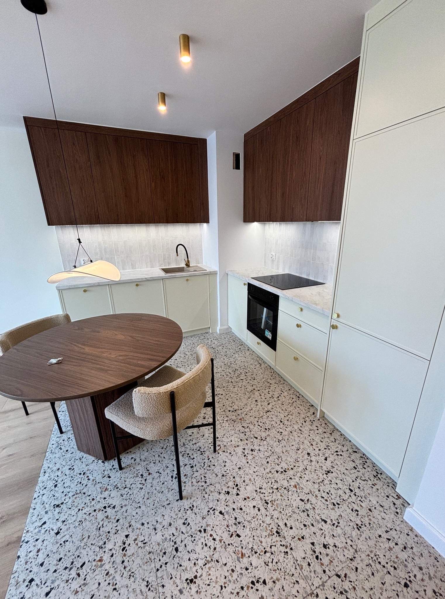 Nowoczesna kuchnia z drewnianymi szafkami wiszącymi, białymi dolnymi szafkami i blatem z marmuru, okrągły stół z ciemnego drewna i krzesła na wzorzystej podłodze.