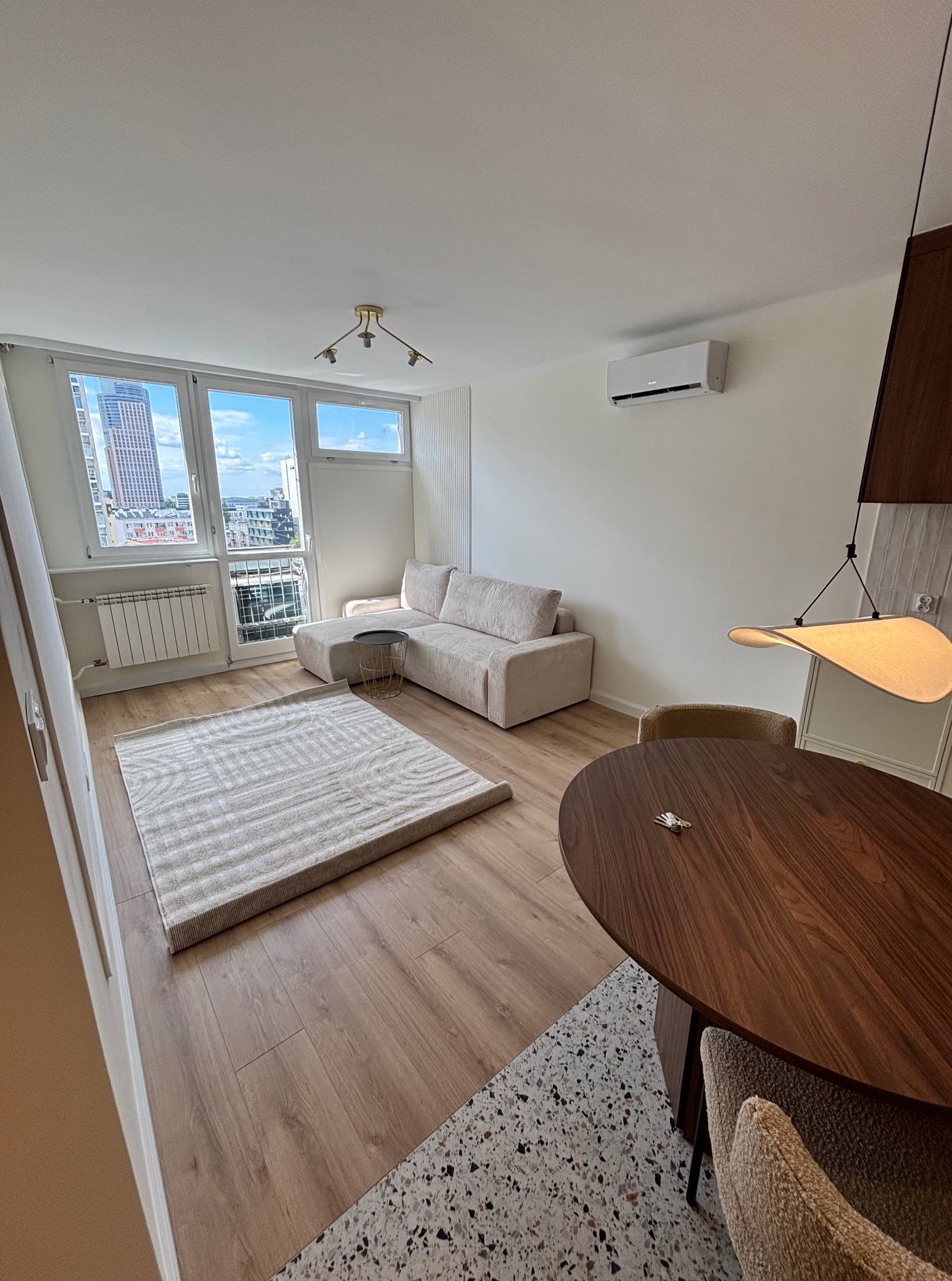 Jasne wnętrze mieszkania z drewnianą podłogą, beżową sofą i okrągłym stołem. Za oknem widok na miasto z wysokimi budynkami.