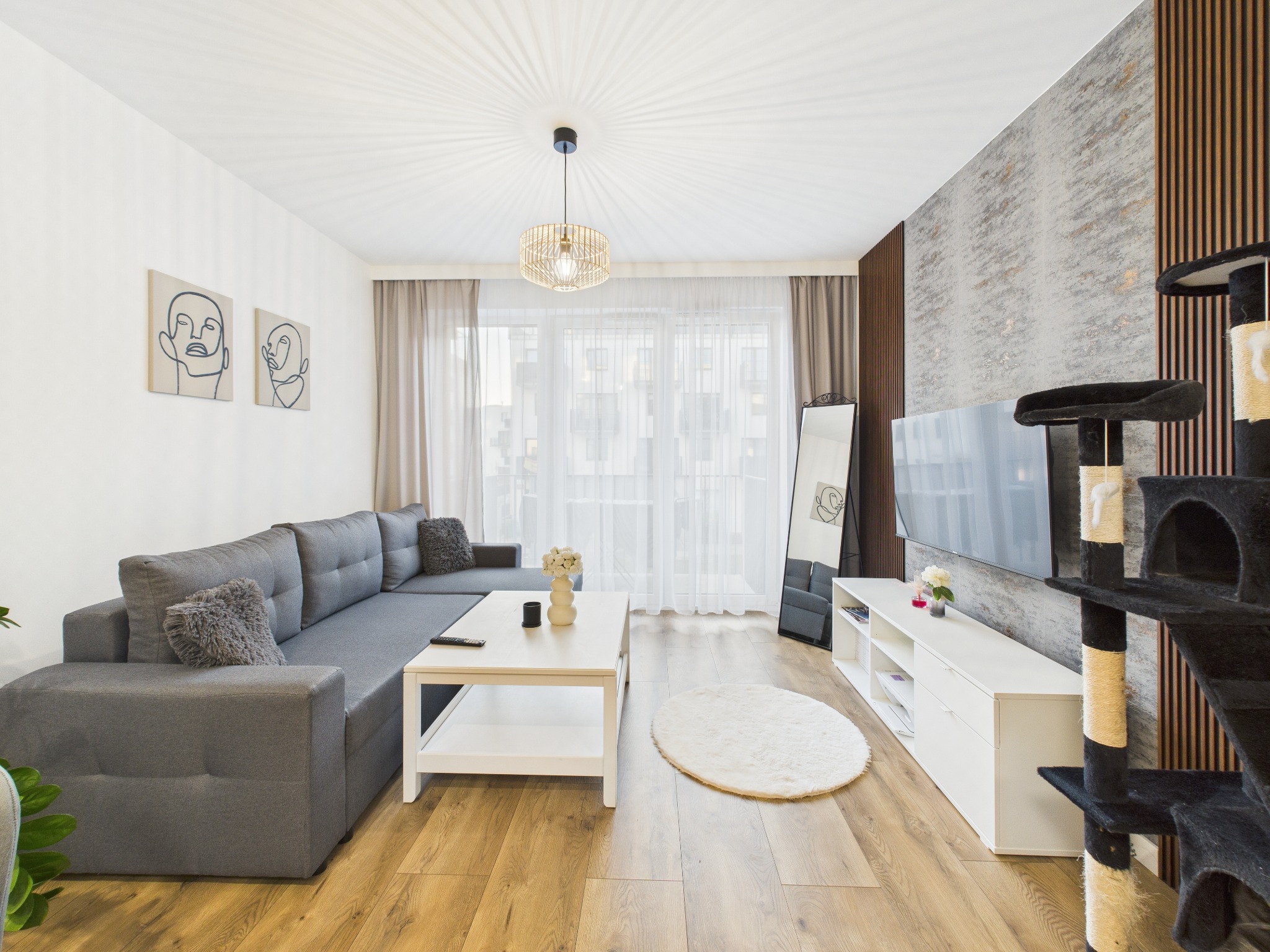 Jasny salon z szarą sofą narożną, białym stolikiem kawowym i drewnianą podłogą. Ściana akcentowa z telewizorem. Lampa sufitowa z wikliny.