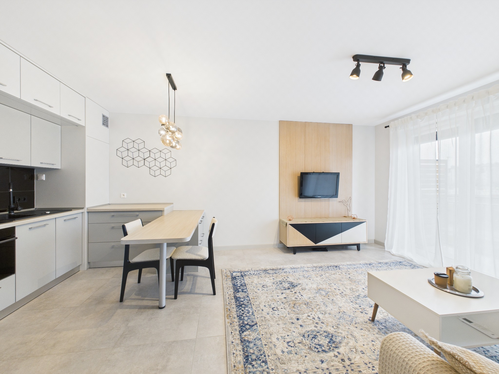 Jasne wnętrze: kuchnia z jadalnią i salonem. Białe meble kuchenne, drewniany stół, dywan w orientalny wzór, telewizor na drewnianym panelu. Nowoczesne oświetlenie.
