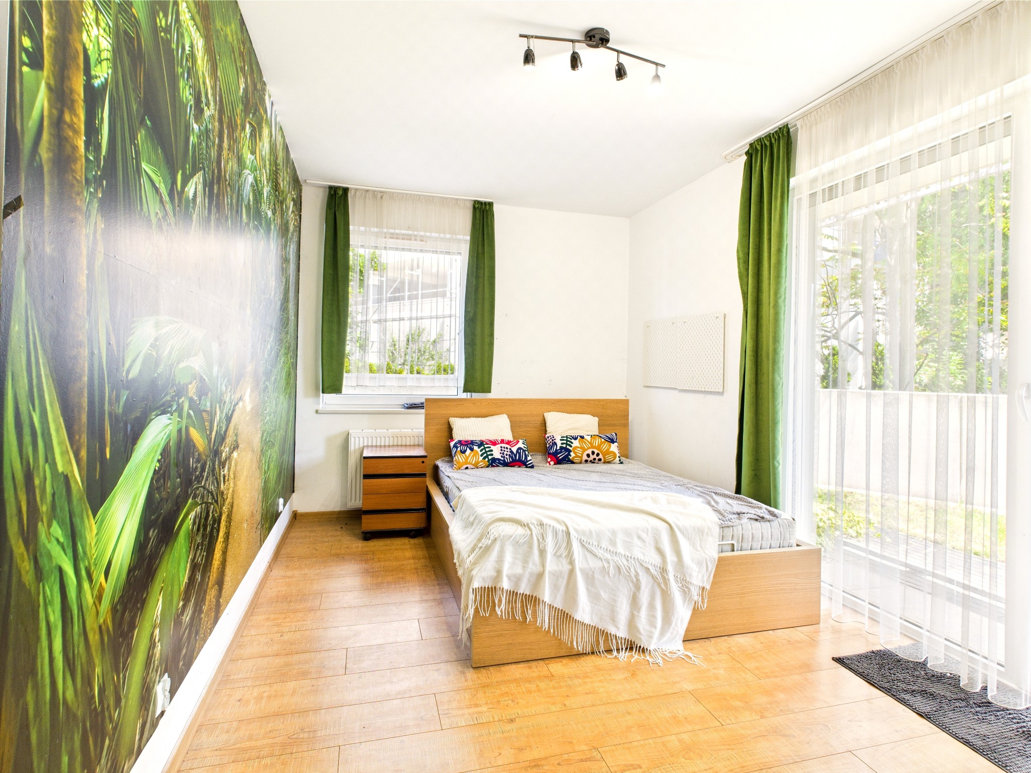 Jasna sypialnia z drewnianą podłogą i łóżkiem. Na ścianie fototapeta z motywem tropikalnym. Okno z zielonymi zasłonami i białymi firankami wpuszcza dużo światła.