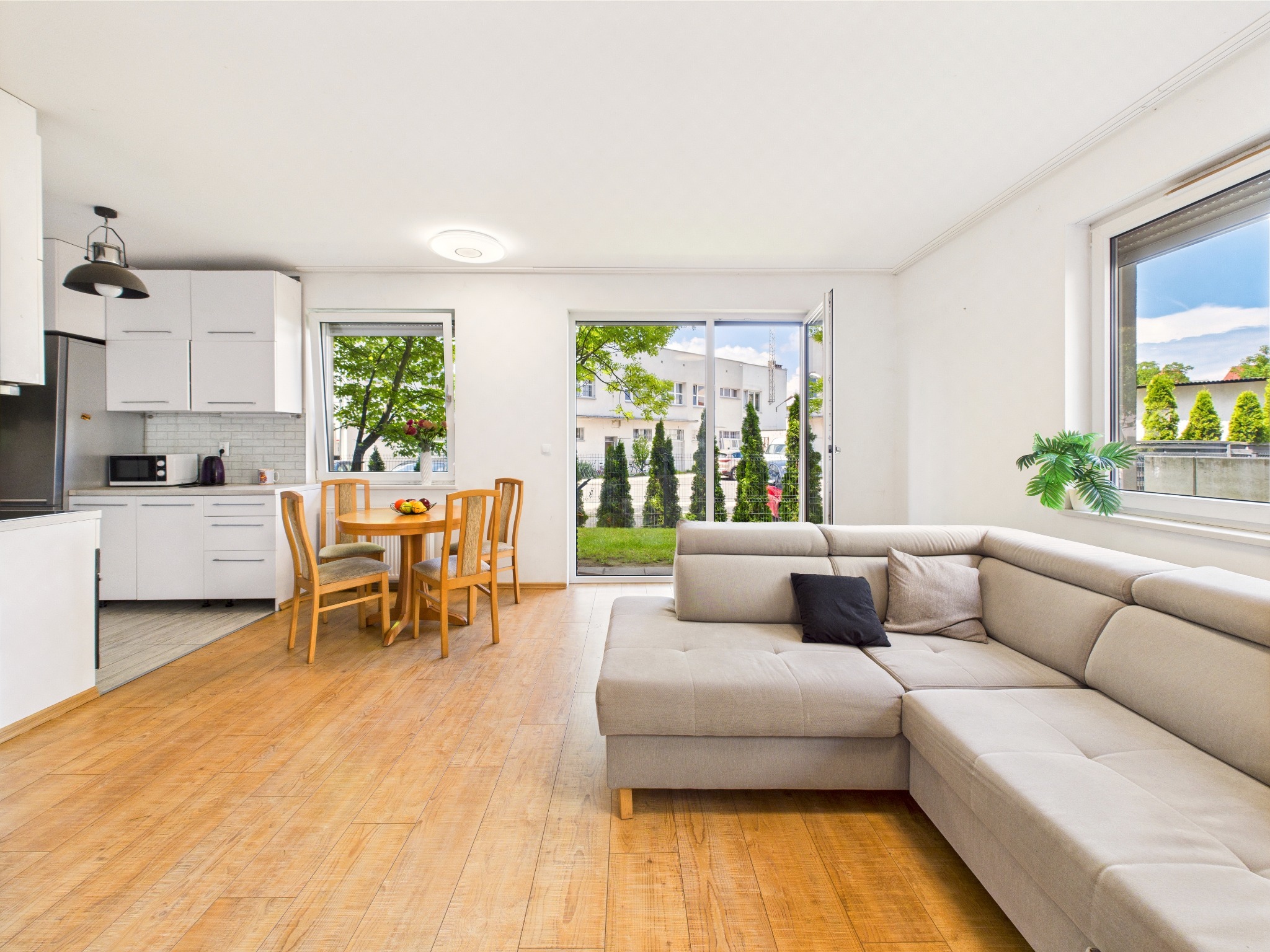 Jasne wnętrze: salon z aneksem kuchennym, drewniana podłoga, szara sofa narożna, okrągły stół z owocami, widok na zieleń przez okna i drzwi balkonowe.