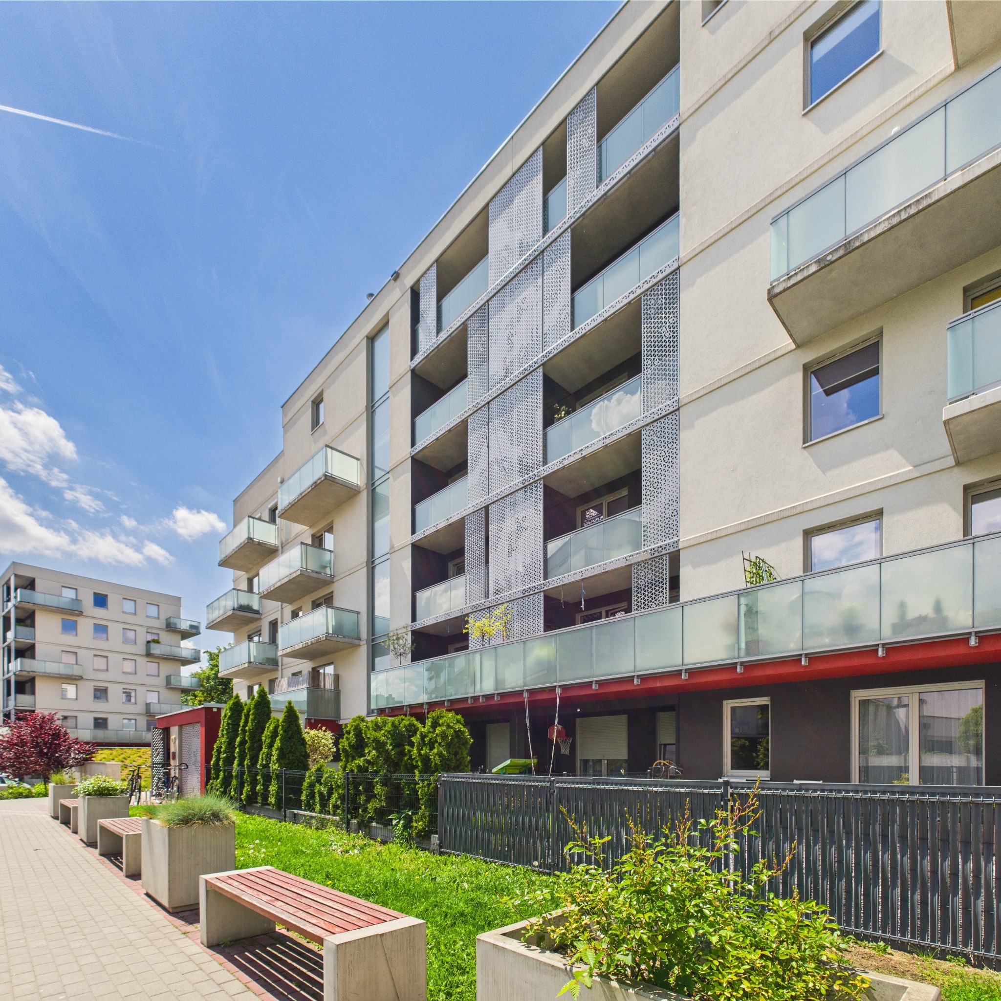 Nowoczesny budynek wielorodzinny z balkonami i ażurowymi panelami. Ujęcie z poziomu chodnika z zielenią i ławkami. Błękitne niebo z delikatnymi chmurami w tle.