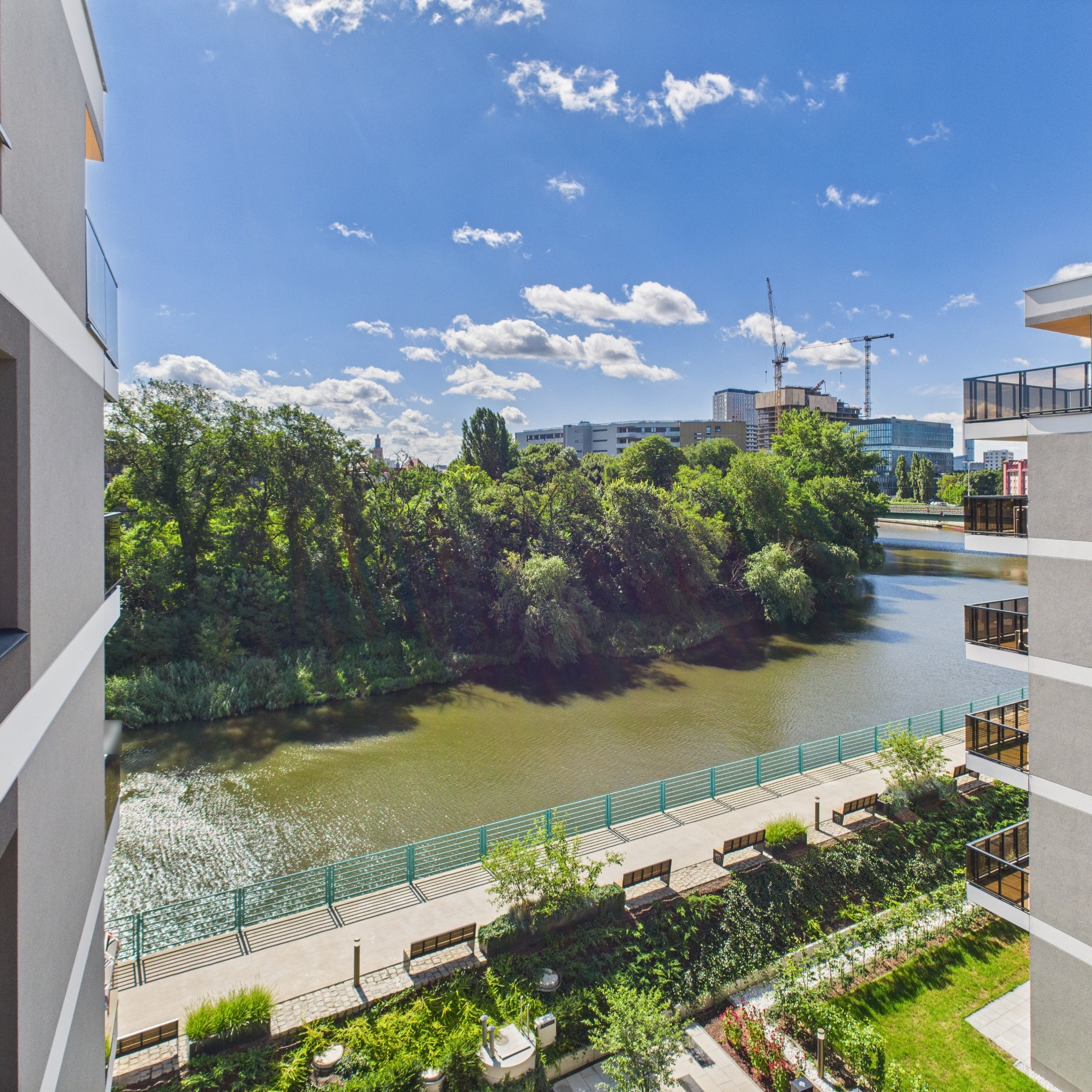 Widok z balkonu na rzekę i park miejski w słoneczny dzień. Nowoczesne budynki po bokach kadru. W tle budowa i dźwigi.